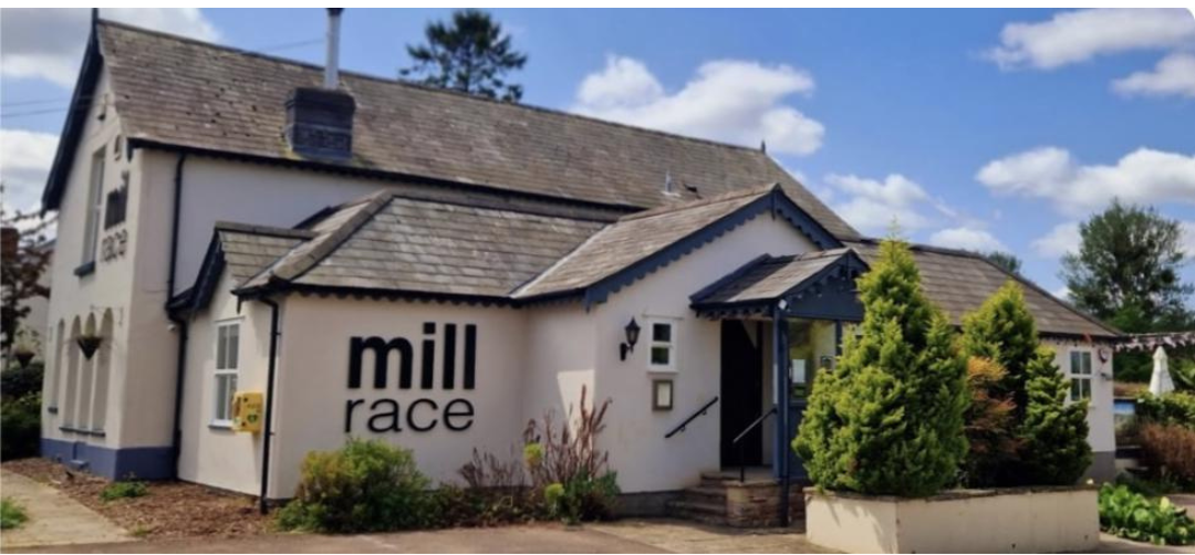 Exterior view of the Mill Race restaurant, a single-story building with white walls and dark roofing. Landscaped with shrubs in the foreground under a sunny sky. A Night to Remember.