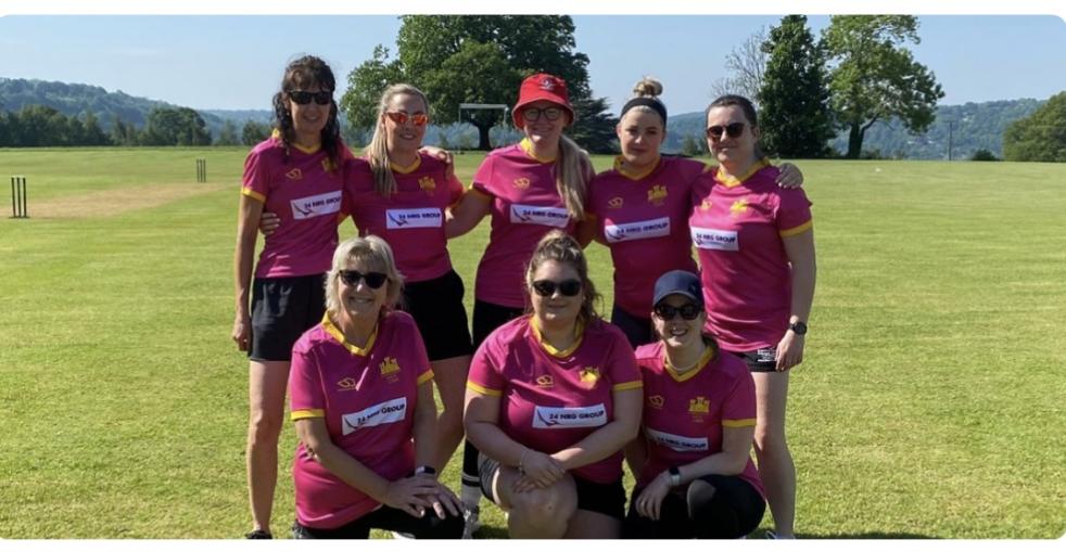 Group of cricket players in pink uniforms standing together on a sunny field, with scenic hills in the background. Goodrich Cricket Club. A Night to Remember.